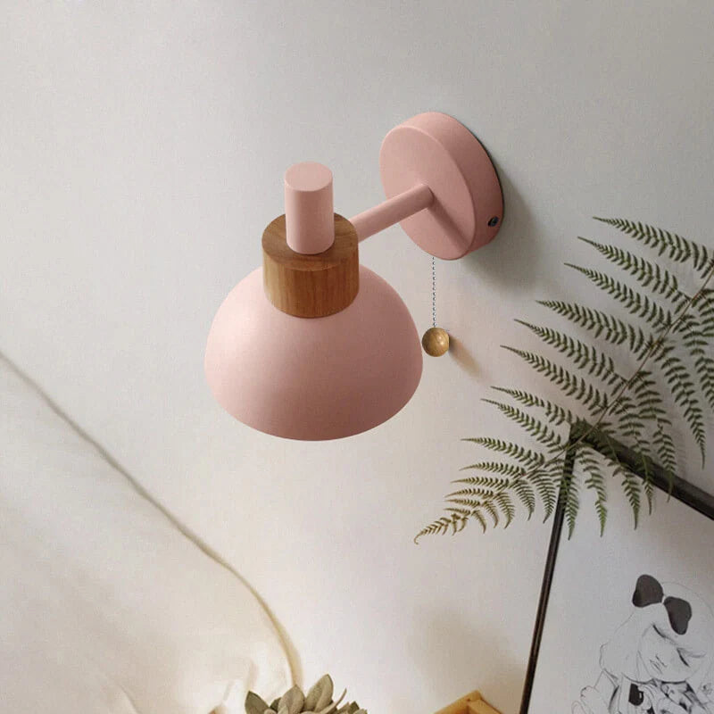 Pink wall lamp with wooden accent on a light gray wall with a plant in the corner.
