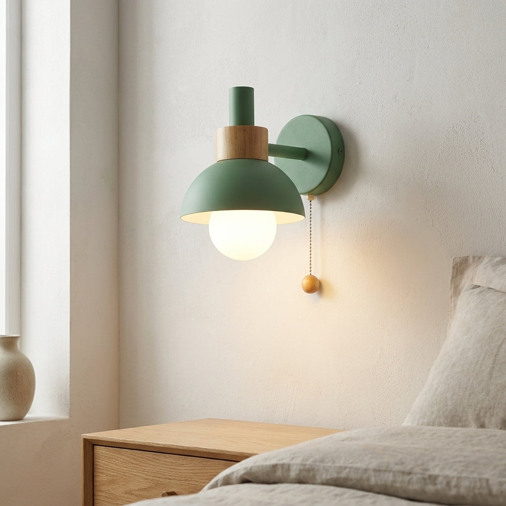 A modern wall-mounted lamp with a sage green dome shade, light wood accent, and glowing bulb, shown installed on a bedroom wall above a wooden nightstand.