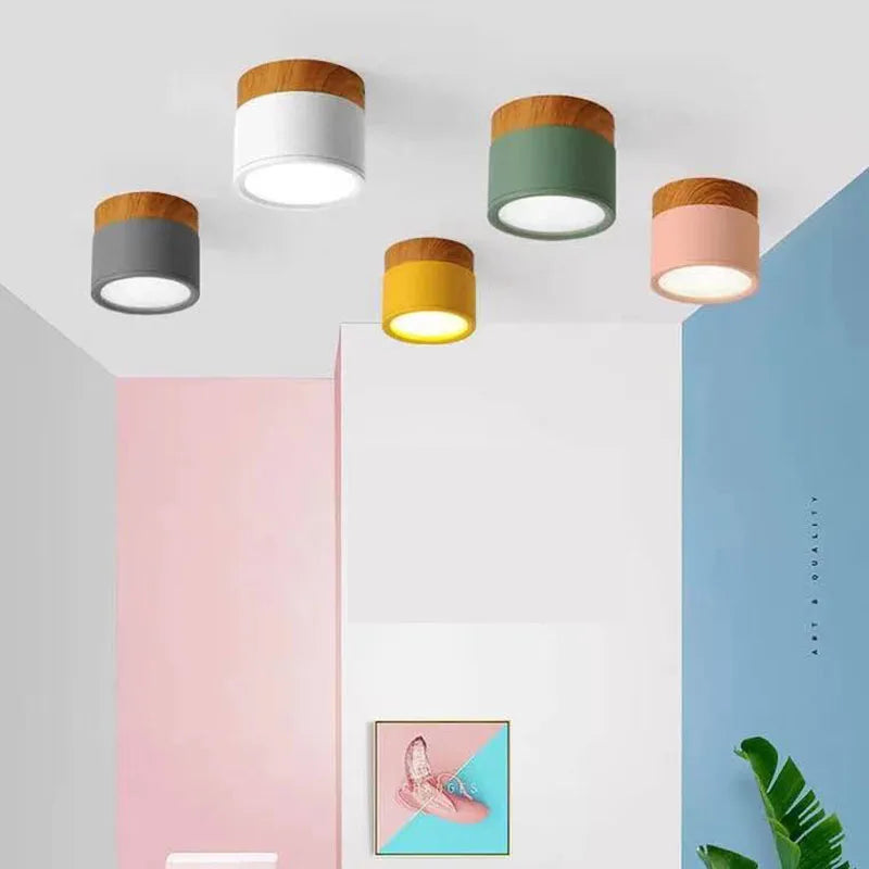 Five modern cylindrical ceiling lights with wood-look caps in gray, white, green, yellow, and pink, shown installed on a white ceiling in a styled room with pastel walls.