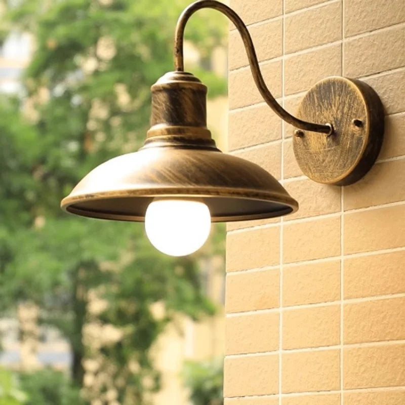 An outdoor wall sconce light fixture with a vintage bronze finish, mounted on a beige tiled wall with a blurred green garden background.