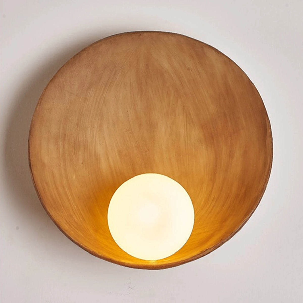 A top-down view of a round wooden bowl or tray with a warm golden-brown finish, featuring a glowing white spherical light bulb placed inside at the bottom center, set against a plain off-white background.
