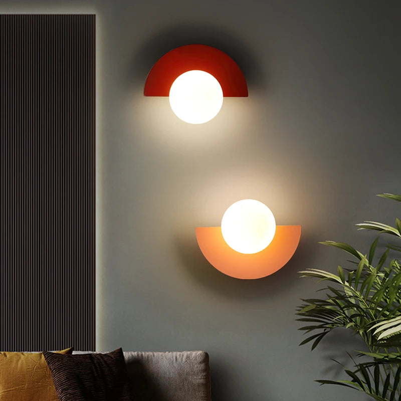Two wall-mounted lights with red and orange light on a gray wall, with a plant and sofa in the foreground.