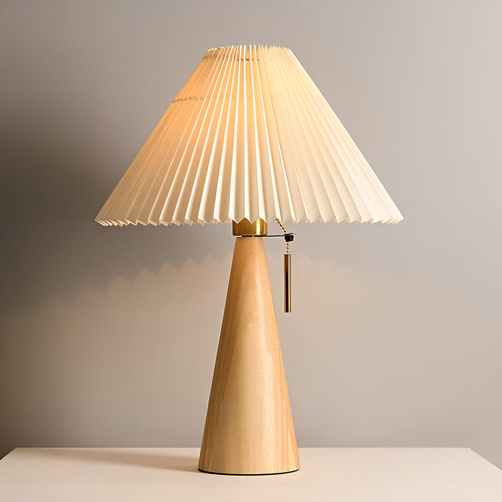 A modern table lamp with a pleated cream fabric shade, a natural wood tapered base, and a brass pull switch, shown on a light surface against a neutral gray background.